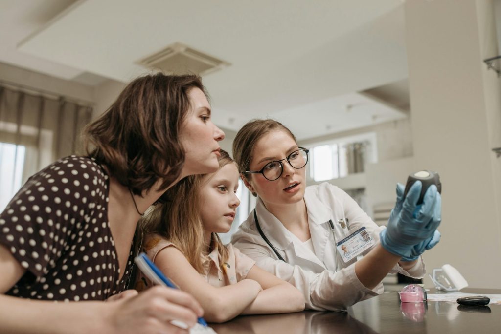 A doctor explains a glucometer to a mother and child in a clinic setting indoors.